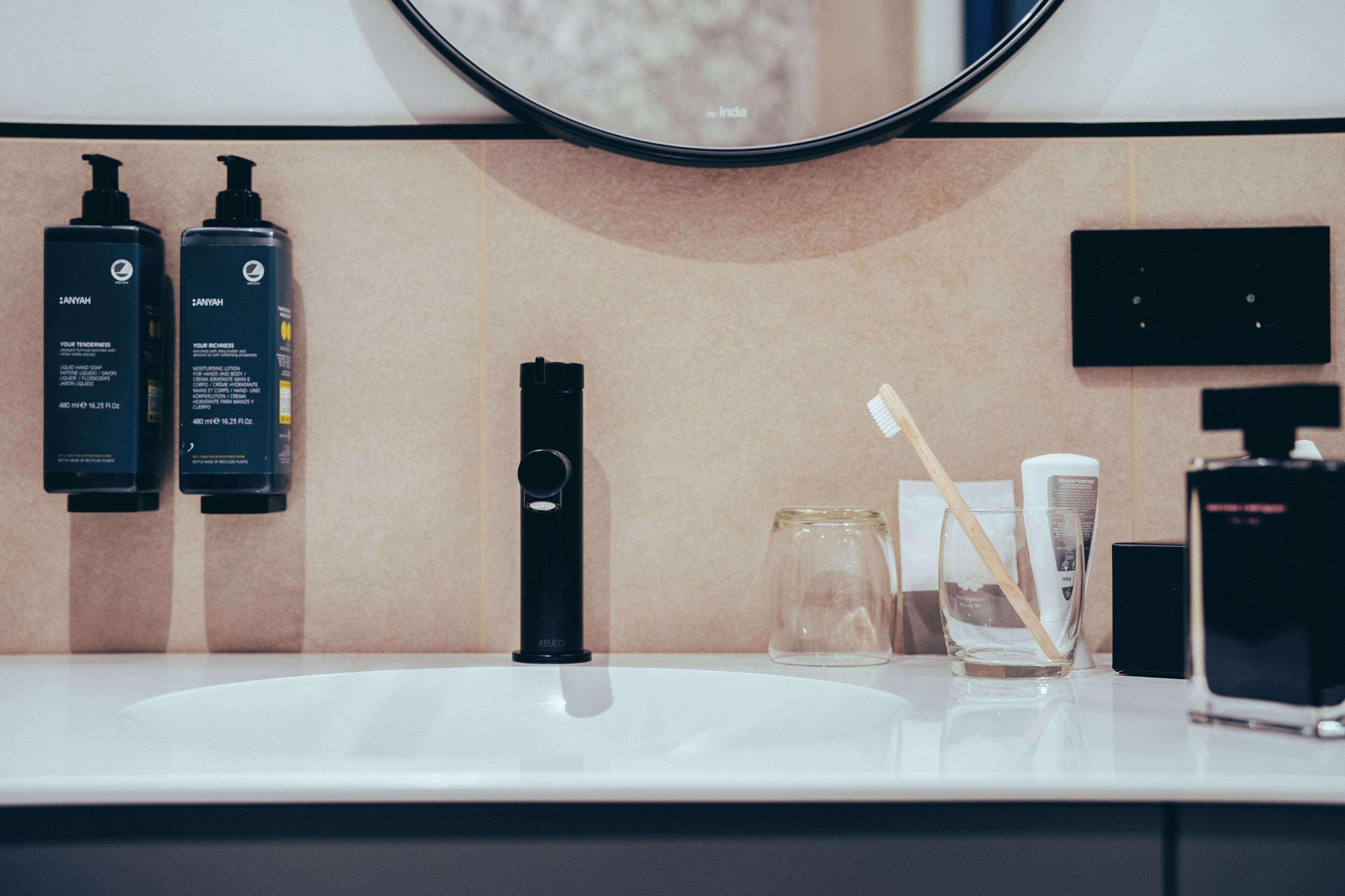 A bathroom sink with a mirror above it, two black bottles of shampoo, a black soap dispenser, a glass with a toothbrush, and a bottle of lotion.
