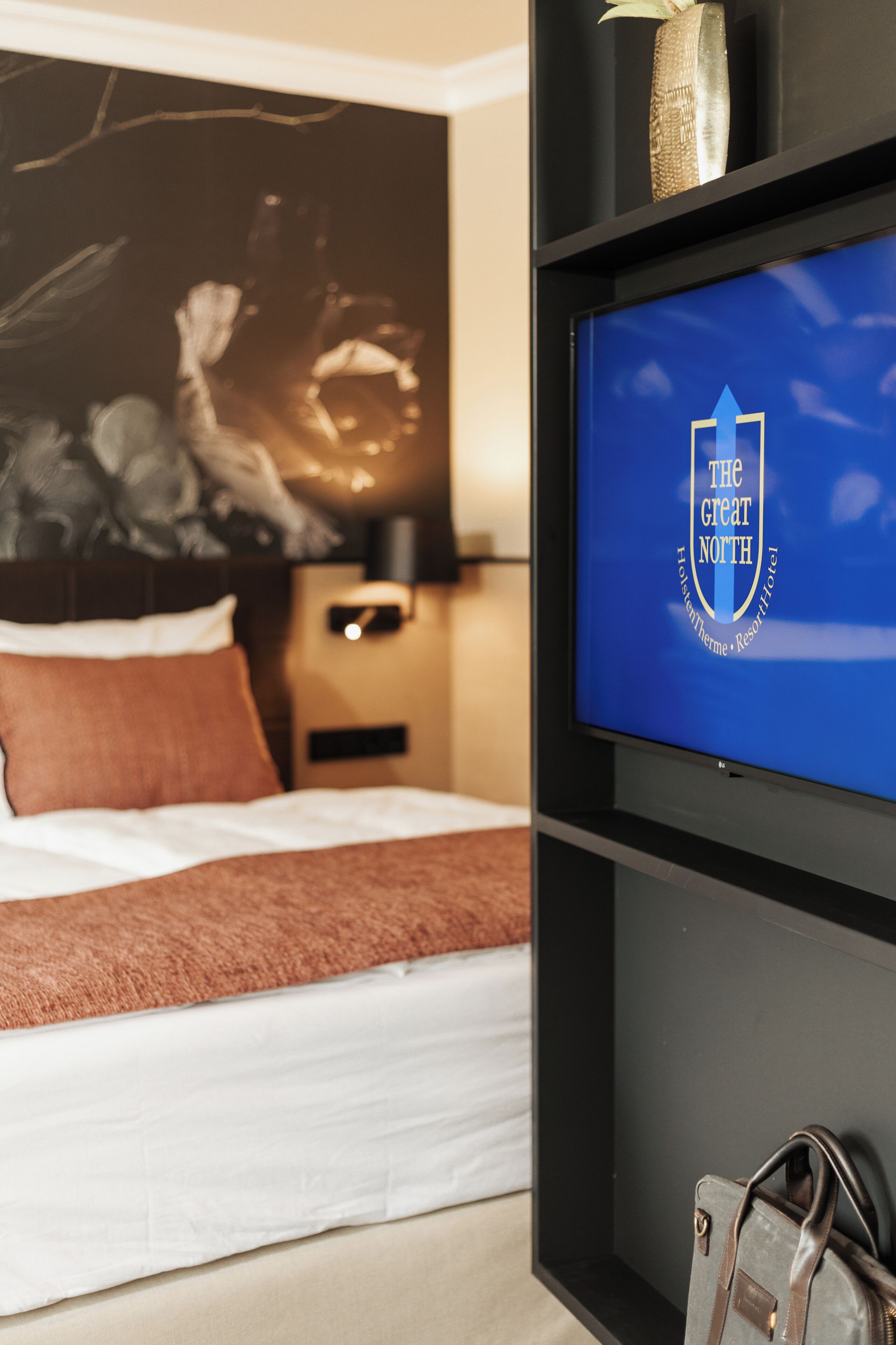 Une chambre d’hôtel avec un lit, un téléviseur affichant le logo de l’hôtel et une table de chevet avec une lampe.