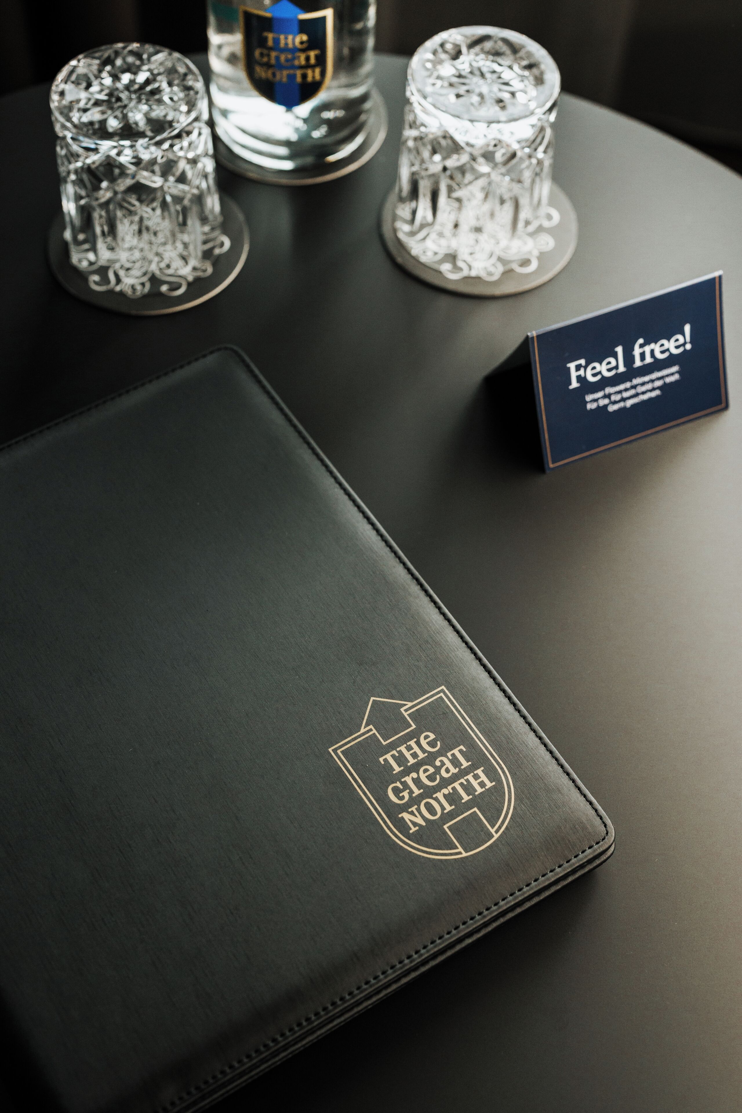 Un cuaderno de tapa de cuero con texto en relieve, dos copas de cristal y una tarjeta sobre una superficie oscura.