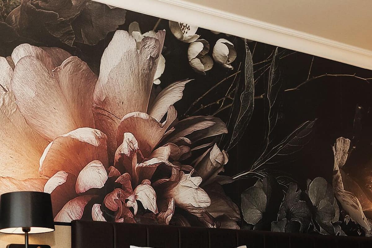 A hotel room with a large floral mural on the wall, a bed with white bedding and multiple pillows, a bedside table with a lamp, and a wooden nightstand.