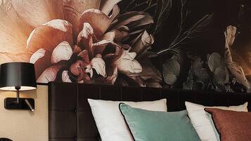 Habitación de hotel con un gran mural floral en la pared, una cama con ropa blanca y varias almohadas, una mesita de noche con lámpara y un tocador de madera.