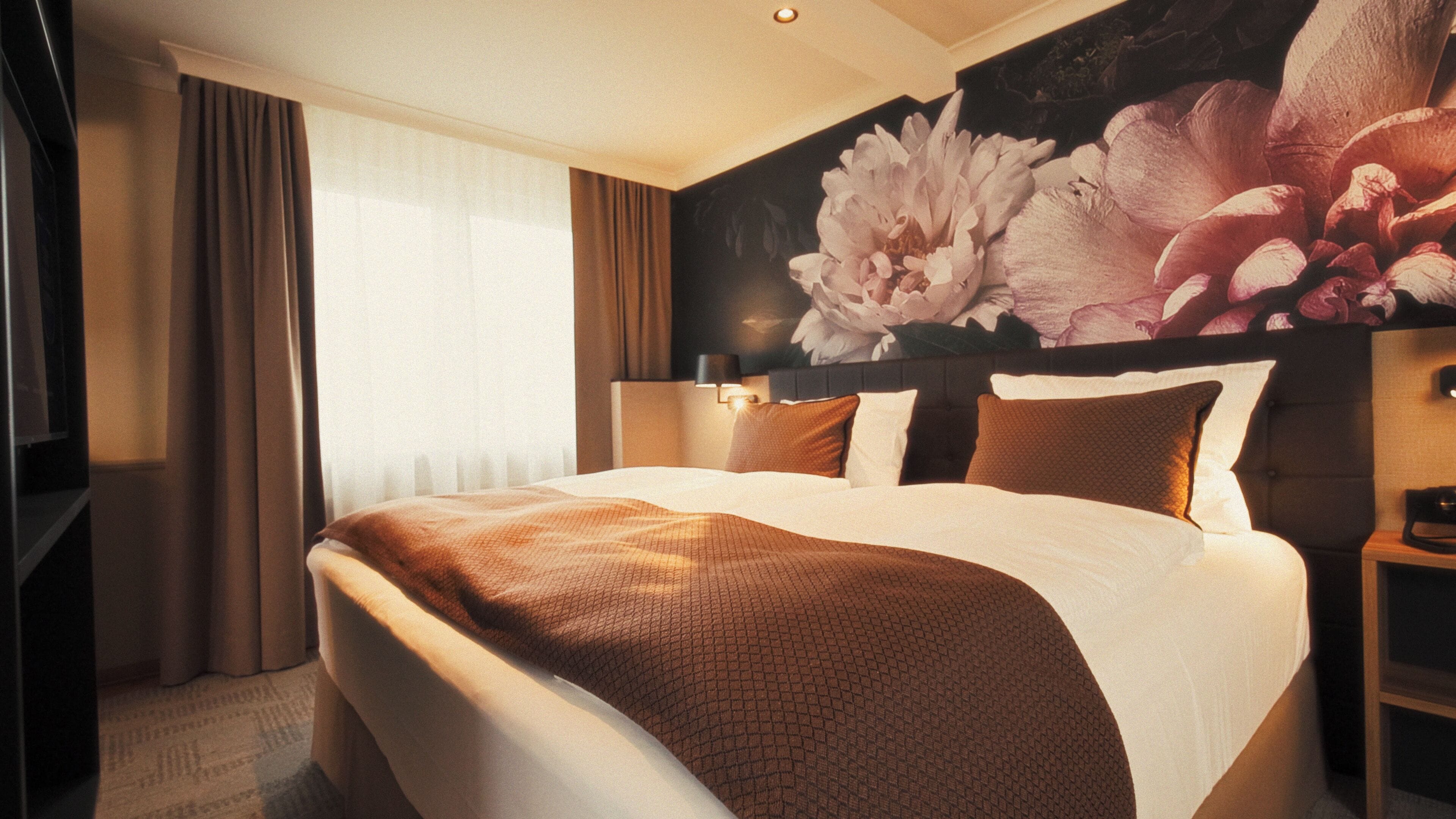 Una habitación de hotel con una cama grande, cabecera con diseño floral, mesita de noche y ventana con cortinas.