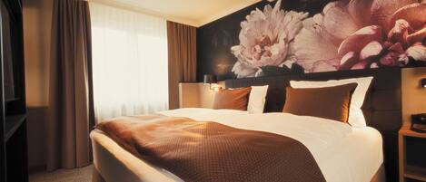 Una habitación de hotel con una cama grande, cabecera con diseño floral, mesita de noche y ventana con cortinas.