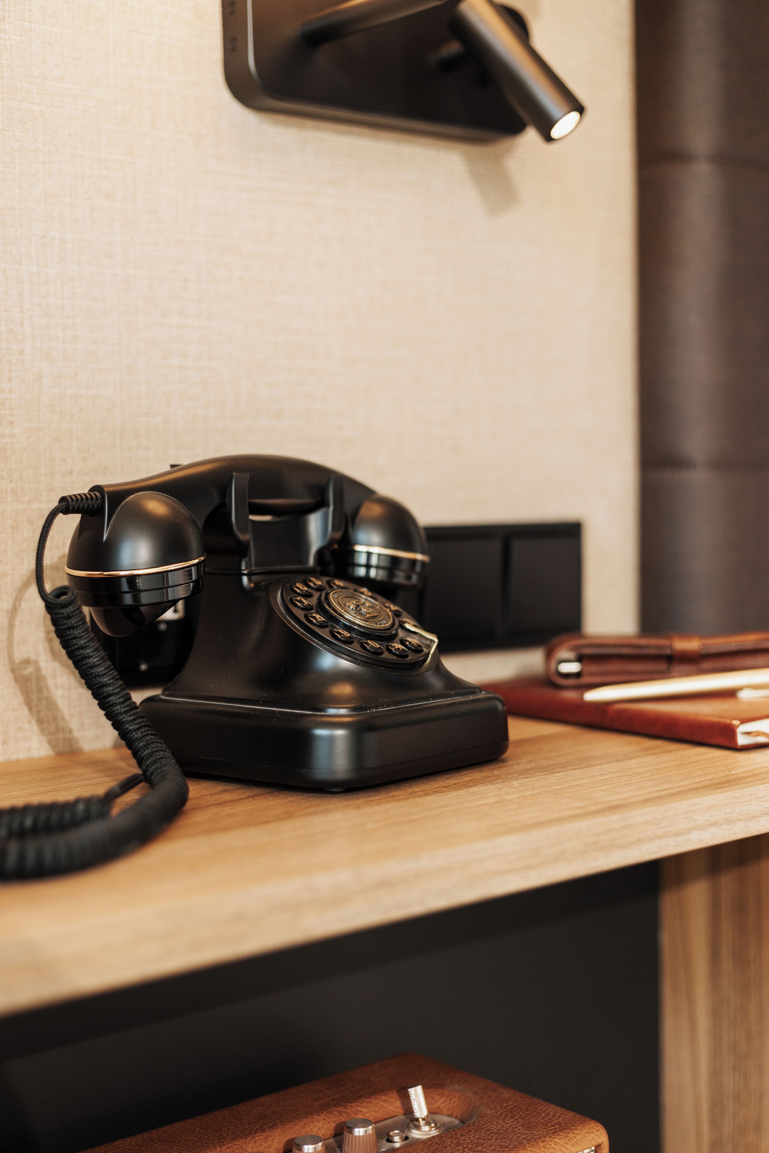 Un téléphone à cadran noir posé sur un bureau en bois, avec une lampe et un livre à la couverture en cuir.