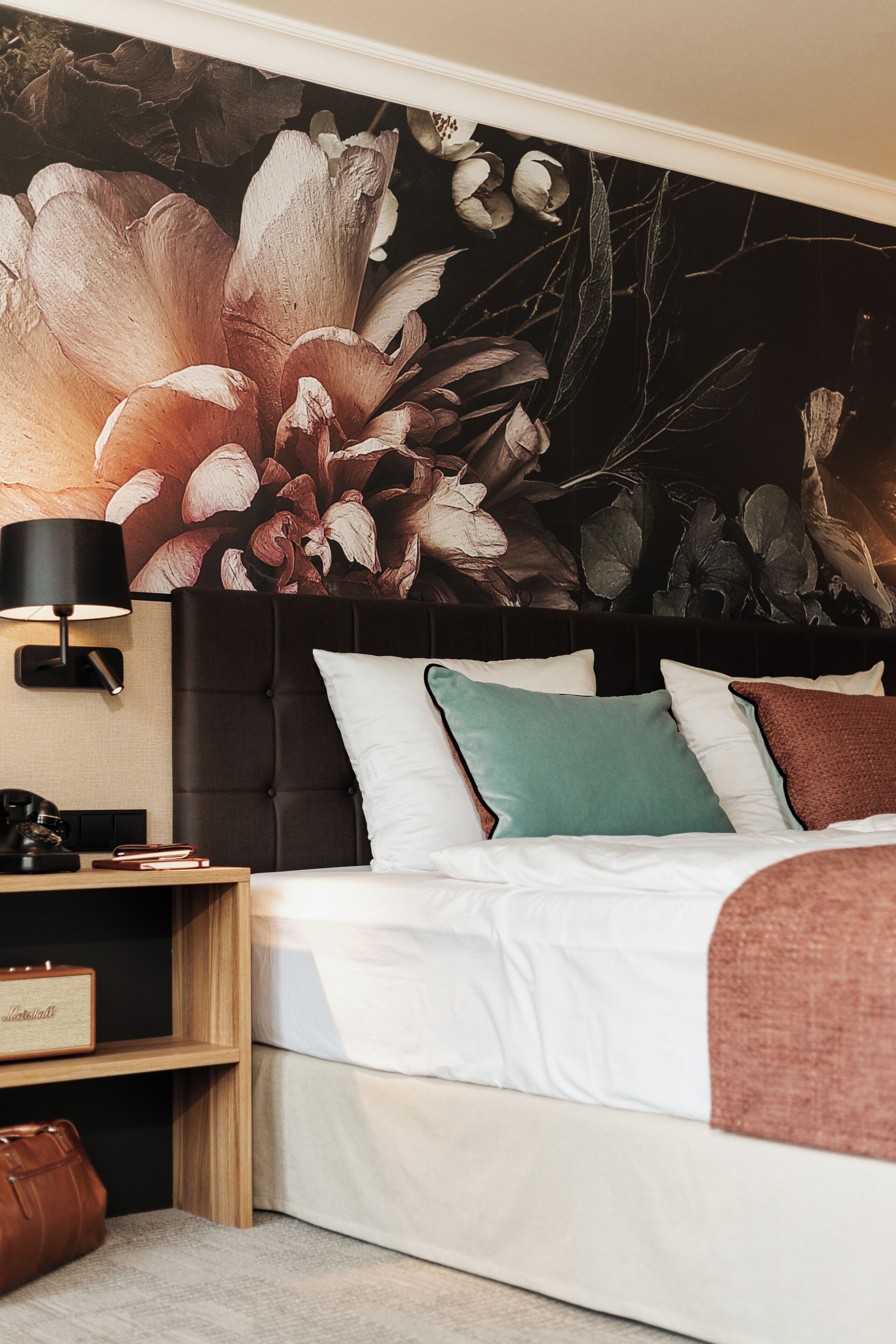 A bedroom with a bed, pillows, a nightstand, a lamp, and a large floral artwork on the wall.