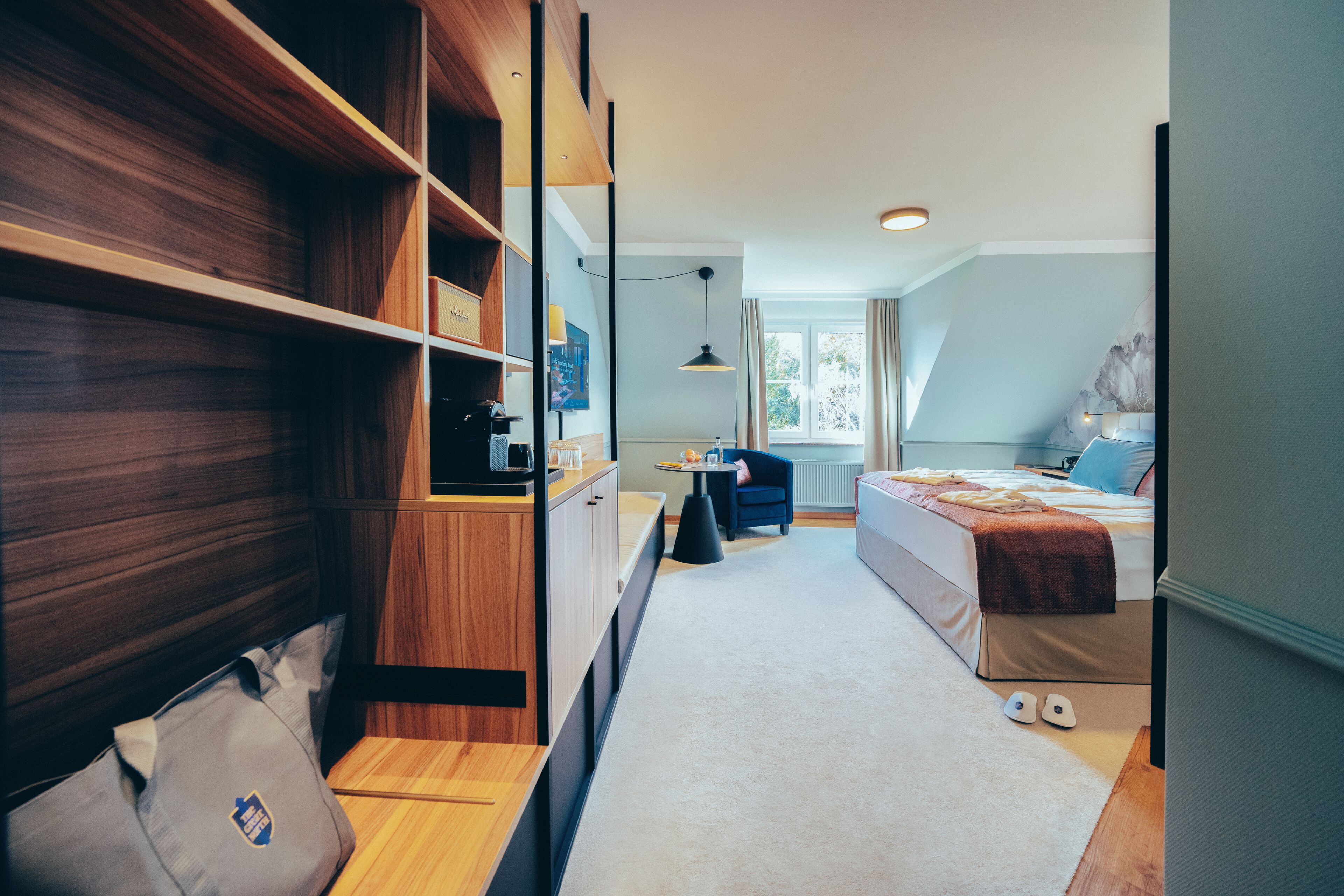 A modern hotel room with a wooden wardrobe, a small desk with a chair, a bed with a blue headboard, and a window with curtains.
