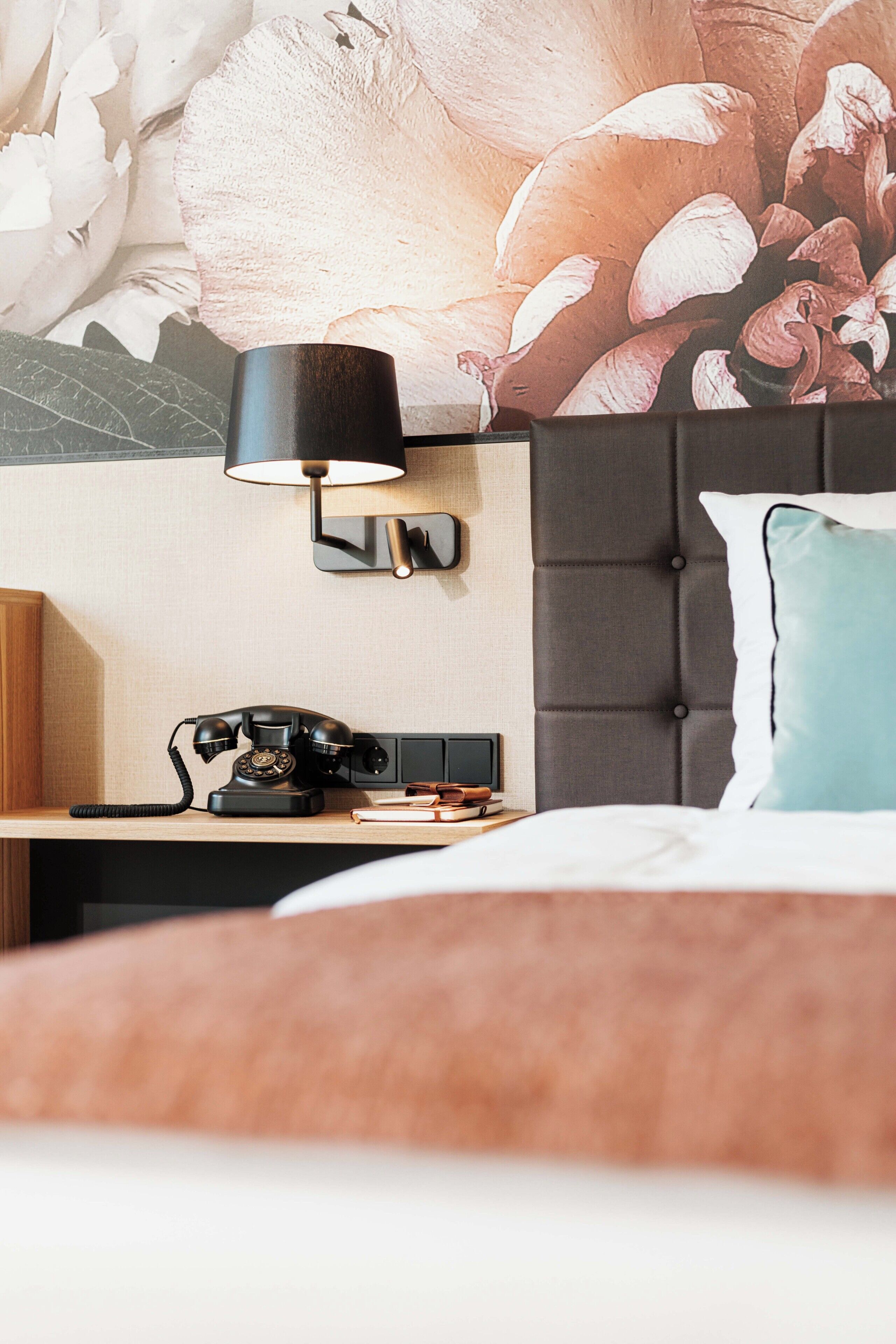 A hotel room with a bed, a nightstand with a telephone, a wall-mounted lamp, and a decorative wall.