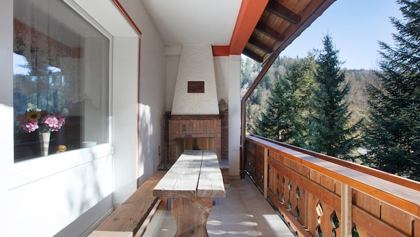 Een balkon met een houten tafel en bankjes, een metselwerk open haard en uitzicht op een bebost helling.
