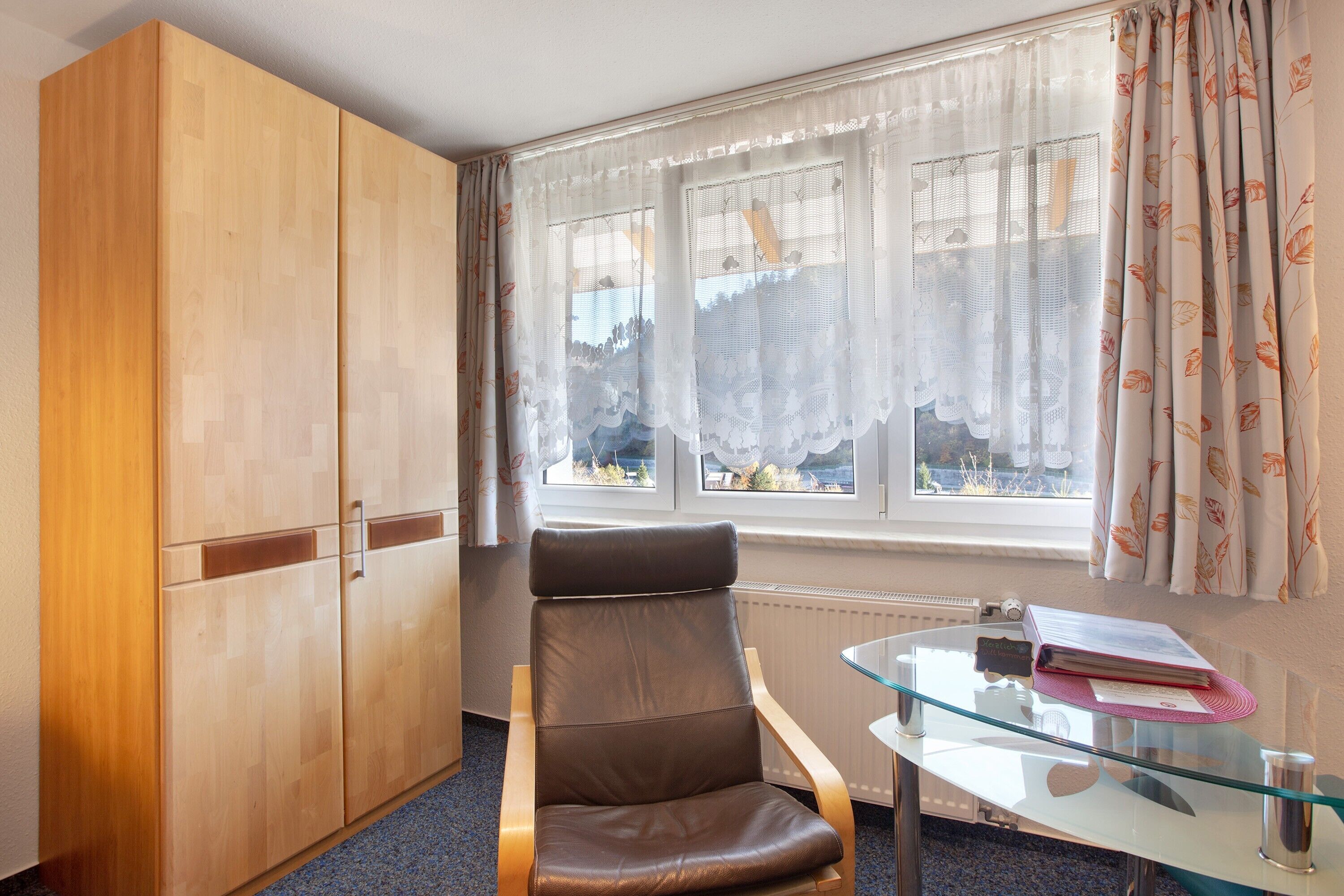 A room with a brown leather chair, a glass table with a book and a pink notebook, and a wooden wardrobe.
