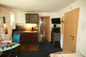 Ein kompaktes Hotelzimmer mit Küchenzeile, einem Einzelbett, einem Schreibtisch mit Stuhl und einem Fernseher.