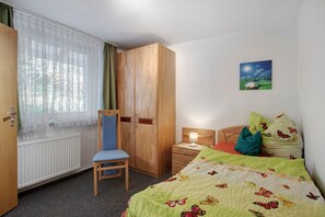 Une chambre à coucher comprenant un lit, une chaise, un bureau, une armoire, un radiateur et une fenêtre avec des rideaux.