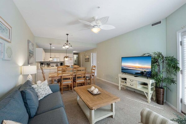 A living room with a sofa, a coffee table, a TV, a dining area with chairs, and a ceiling fan.