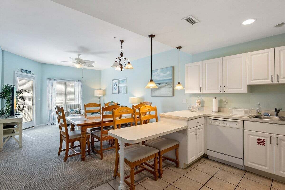 Un área de comedor con una mesa y sillas, ventilador de techo, lámparas colgantes y una cocina con armarios blancos y lavavajillas.