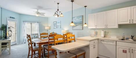 Un área de comedor con una mesa y sillas, ventilador de techo, lámparas colgantes y una cocina con armarios blancos y lavavajillas.