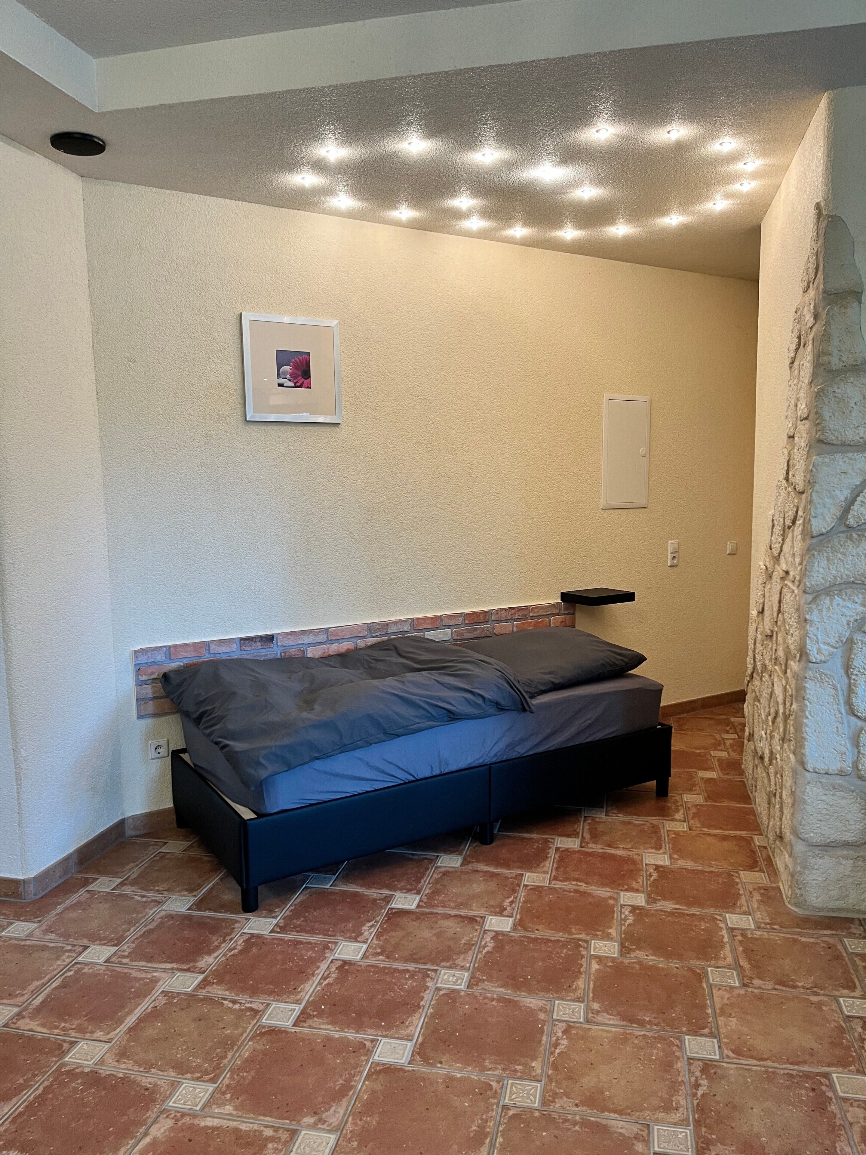 Ein Schlafzimmer mit Ziegelmuster-Kopfstütze, einem Bett mit dunkler Bettwäsche, einem gerahmten Bild an der Wand und einer Steinwand an der Seite.