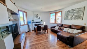 Ein modernes Wohnzimmer mit einem braunen Ledersofa, Holzboden, einem Esstisch mit Stühlen, einer Küchenzeile im Hintergrund und großen Fenstern, die für natürliches Licht sorgen.