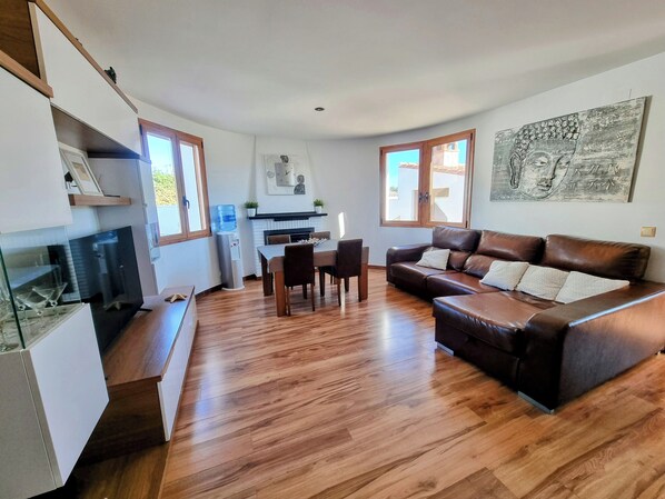 Una sala de estar moderna con un sofá de cuero marrón, piso de madera, una mesa de comedor con sillas, un área de cocina al fondo y ventanas amplias que dejan entrar luz natural.
