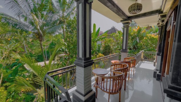 A balcony with wooden chairs and a table, surrounded by lush greenery and a view of a traditional building. - Surya Ubud View and Suites by EPS (Tegallalang)