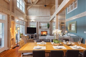 A spacious living room with a fireplace, wooden beams, a large TV, and a dining area set for six.