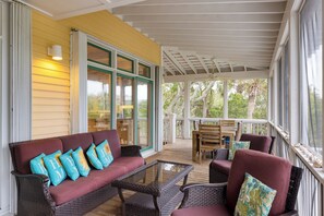A covered porch with a wicker sofa, a glass coffee table, and a wooden table with chairs.