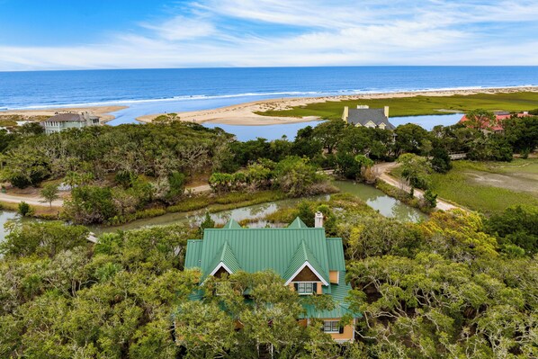 A coastal property with a green-roofed house surrounded by dense greenery, a small pond, and a beach in the background.