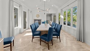 Un vaste espace dédié à la salle à manger, avec une longue table et des chaises à l’assise rembourrée de tissu bleu, de grandes fenêtres ornées de rideaux et un lustre moderne.