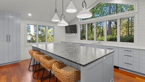 Une cuisine moderne dotée d’un grand îlot central, de tabourets de bar et de grandes fenêtres offrant une vue sur les arbres.