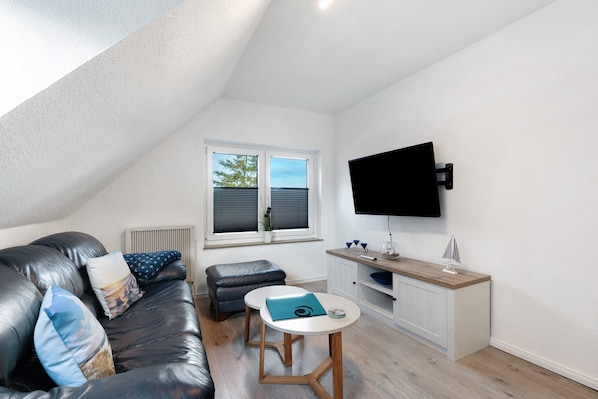 Una sala de estar moderna con un sofá de cuero negro, una mesita redonda pequeña, un mueble blanco y un televisor montado en la pared.
