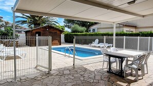 A covered outdoor area with a swimming pool, stone flooring, white furniture, and a wooden cabin.