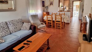 A living room with a sofa, armchair, wooden dining table, and chairs. There is a kitchen area visible in the background.