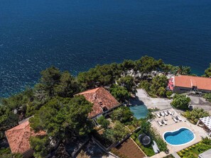 Vue aérienne d’une propriété située sur la côte, avec une piscine, un espace de détente et des bâtiments aux toits rouges, le tout entouré d’arbres.