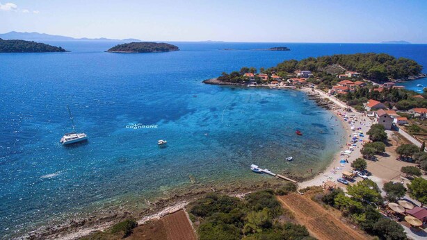 Une vue aérienne d’une ville côtière, avec une eau d’un bleu limpide, des bateaux et une plage parsemée de parasols et de personnes.