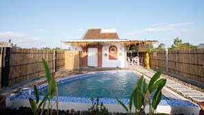 Una piscina con borde de azulejos, una cerca de madera y un edificio con techo de paja al fondo.