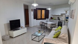 Una sala moderna con una televisión de pantalla plana, un sofá con cojines verdes, una mesa de centro y una zona de comedor con mesa y sillas.