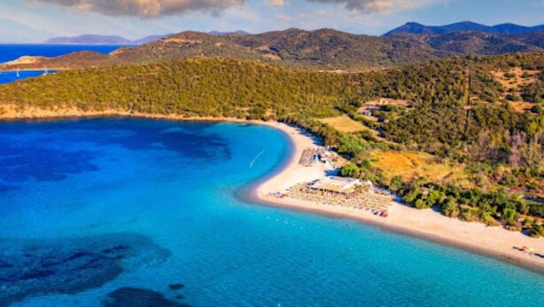 Vista aérea de una zona costera con playa, aguas azul turquesa y montañas al fondo.