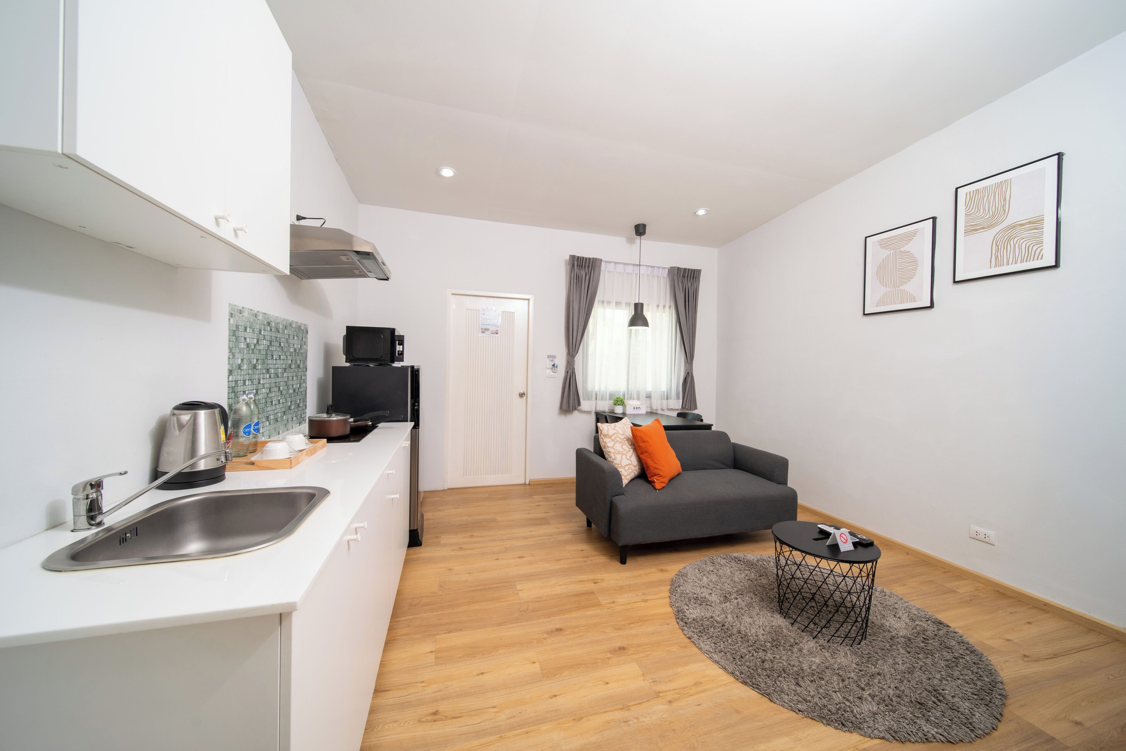 A modern kitchen with a sink, a coffee maker, and a television. There is a grey sofa with an orange pillow in the living area, and a round rug on the floor.