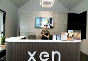 Una reception con un grande schermo sopra, una persona dietro al banco e diversi manifesti informativi e un vaso di fiori sul bancone.