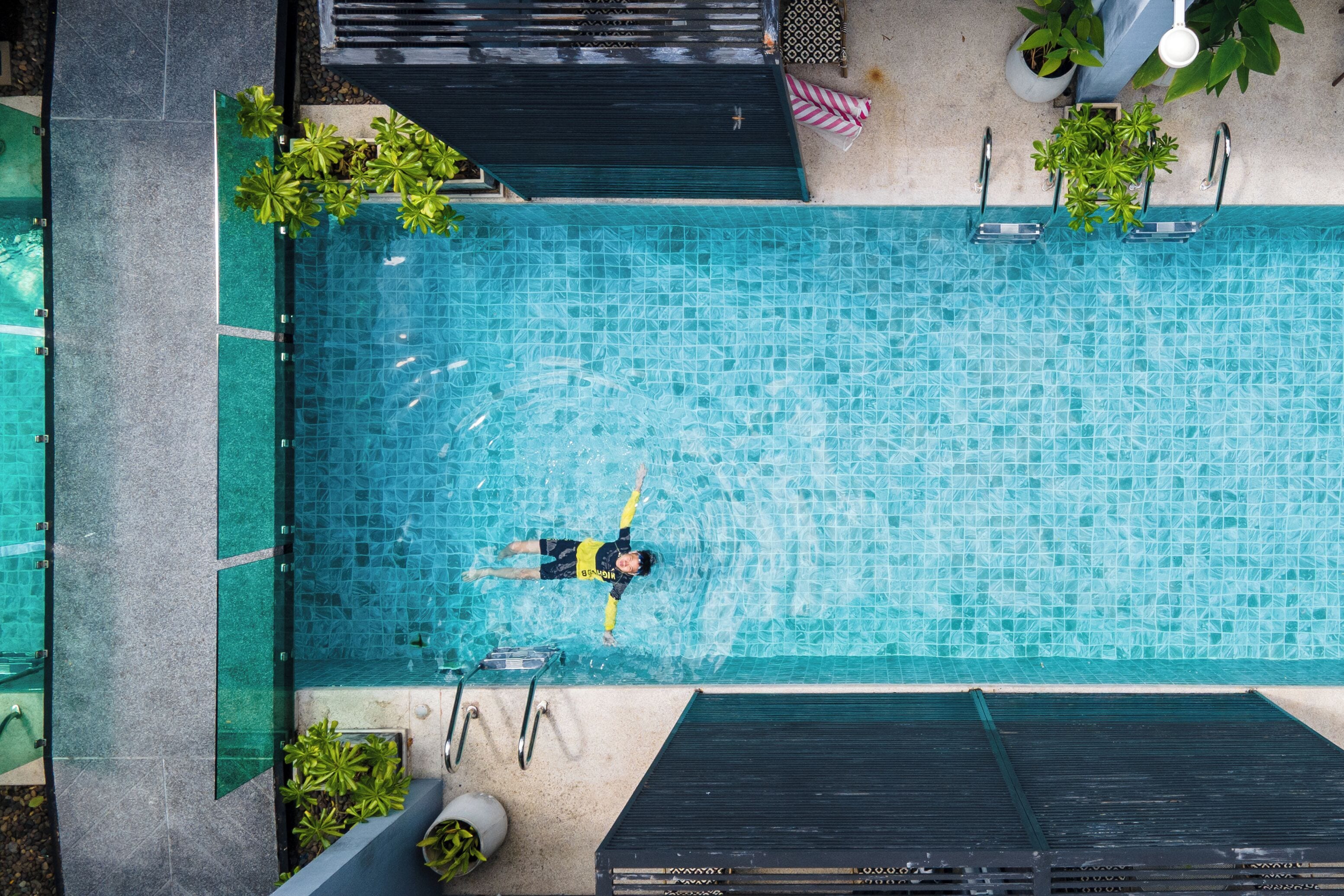 Ein rechteckiger Pool mit Fliesen, umgeben von einer Betonplattform, Topfpflanzen und einer Metallleiter.