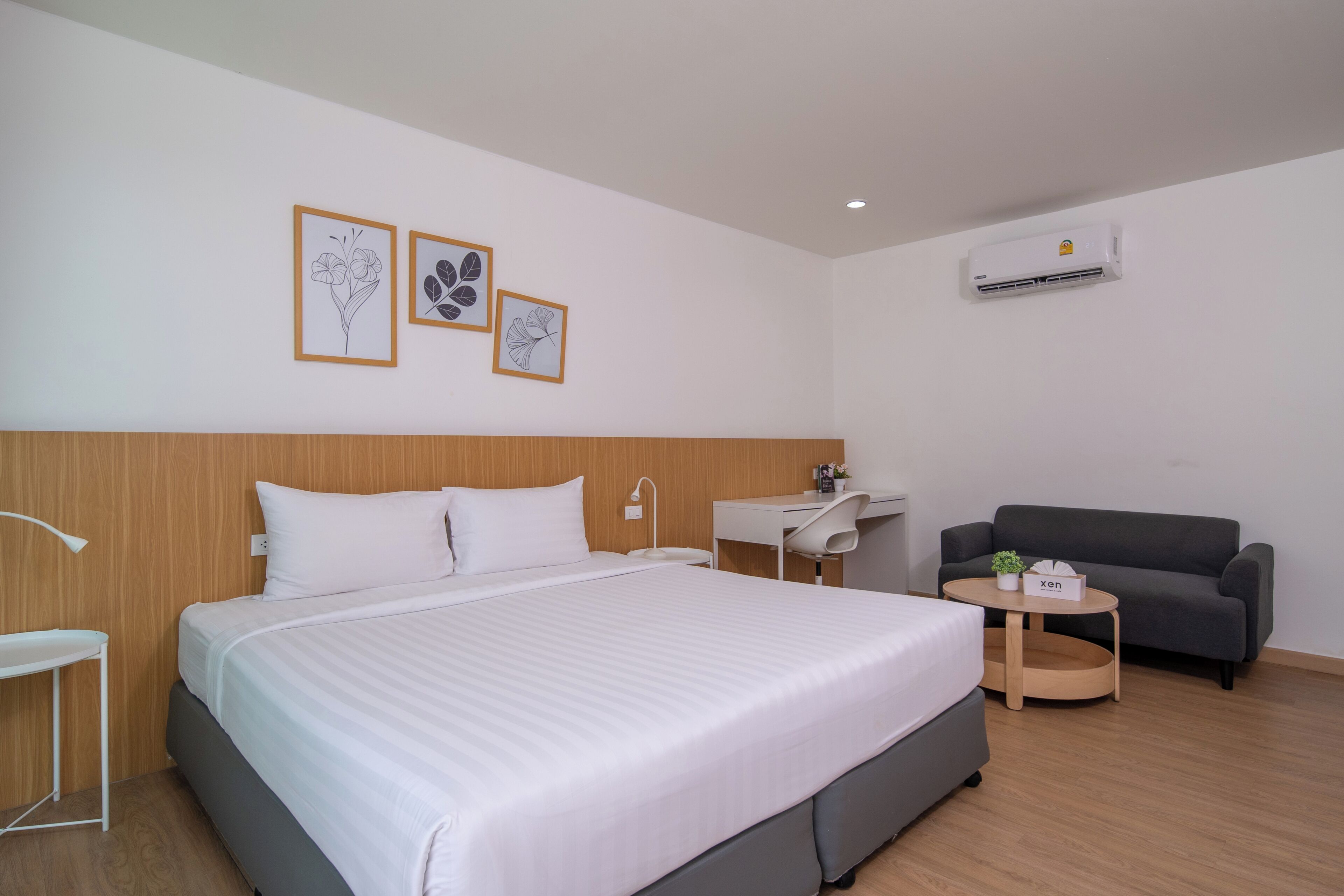 A hotel room with a bed, a desk, a chair, a sofa, and wall-mounted air conditioning.