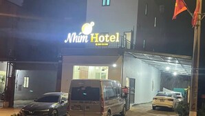 Un hotel llamado "Nhın Hotel" con un diseño moderno, que cuenta con un estacionamiento y varios vehículos, incluyendo una furgoneta y un coche.