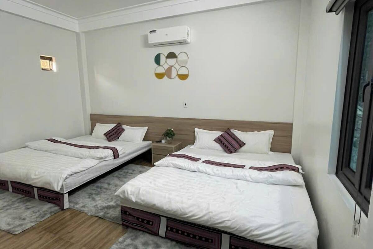 Ein Hotelzimmer mit zwei Betten, einem grauen Teppich, Holzböden, einer Wandklimaanlage und einem kleinen Fenster.