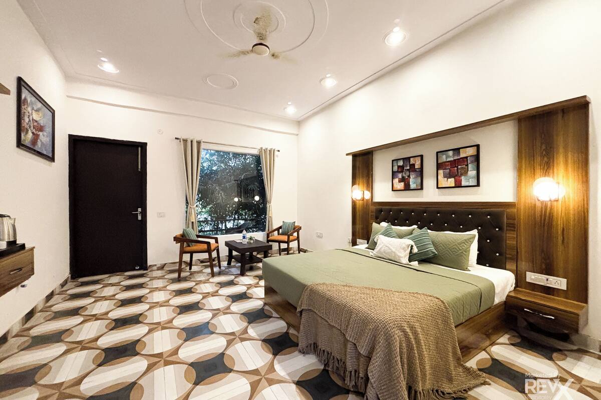 A hotel room with a bed, a desk, two chairs, a window with curtains, a ceiling fan, and a patterned floor.