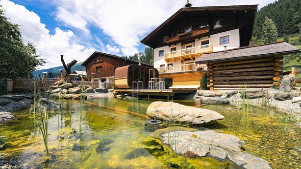 Ein traditionelles Holzchalet mit Balkon, ein klares Teichgewässer mit Steinen und eine Bergkulisse im Hintergrund.
