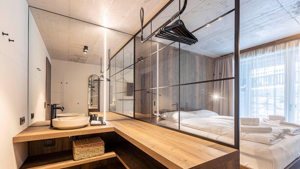 Moderni hotellihuone, jossa on puinen meikkipöytä, suuri peili, valkoisilla lakanoilla varustettu sänky ja suuret ikkunat, joista avautuu näkymä kaupunkiin.