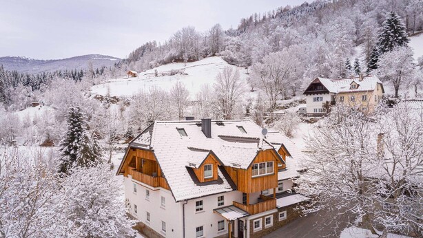 Ein mehrstöckiges Gebäude mit geneigtem Dach, umgeben von schneebedeckten Bäumen und einer hügeligen Landschaft.