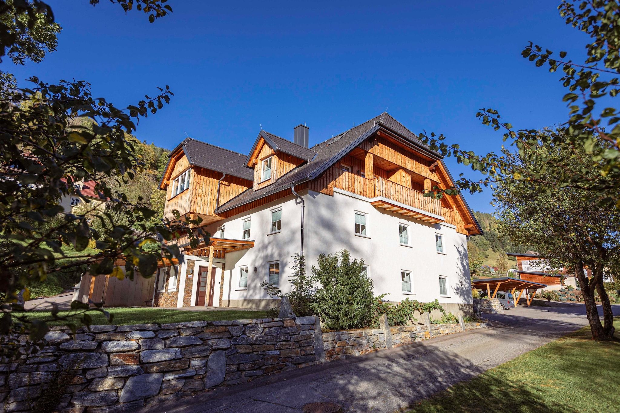 A large, two-story wooden house with a stone wall in front, surrounded by trees and a clear blue sky.