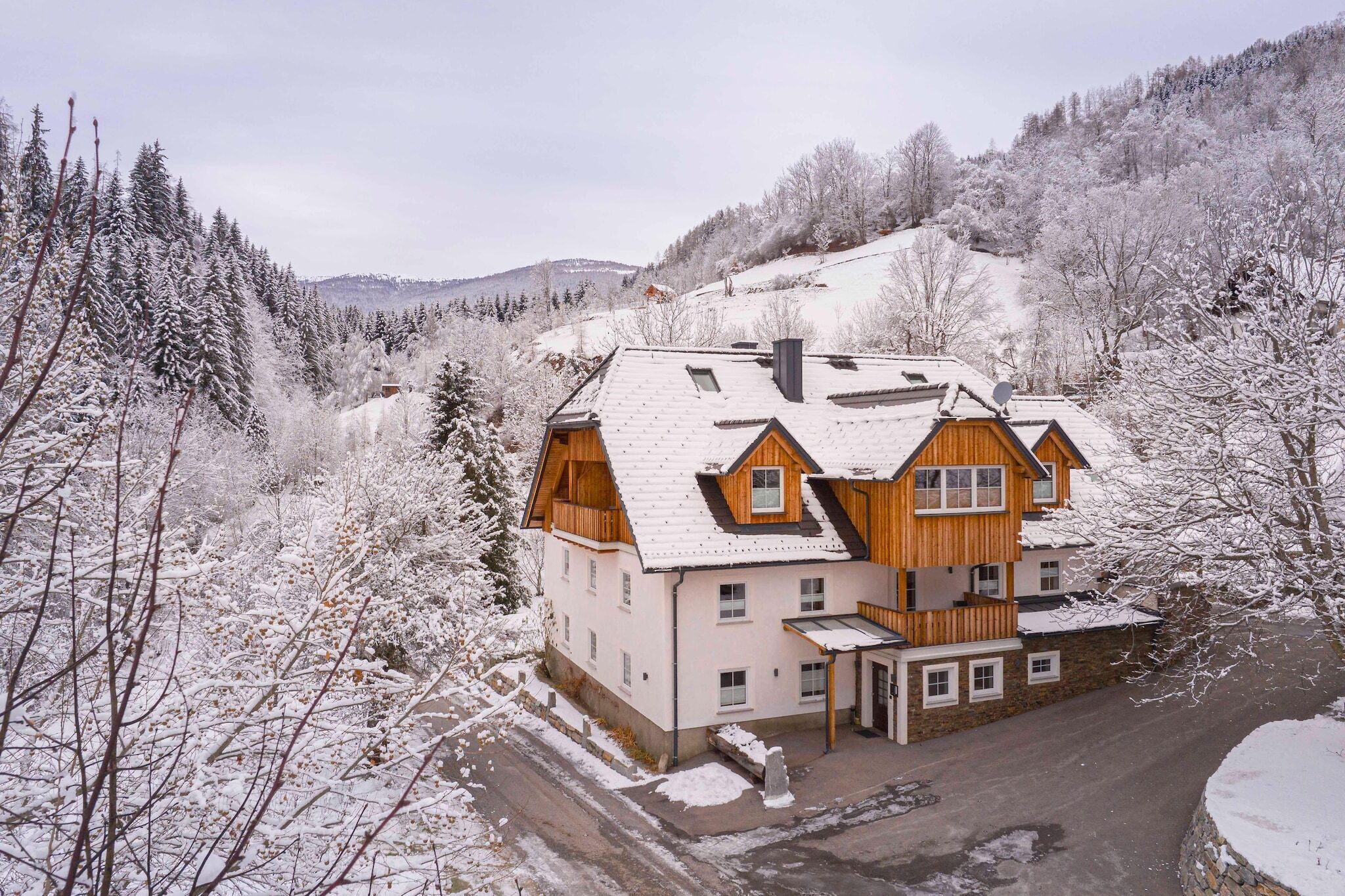 A two-story wooden house with a sloped roof, surrounded by snow-covered trees and hills.