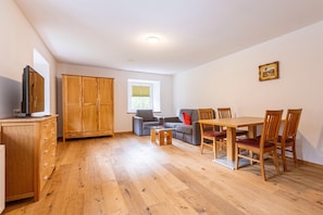 Une pièce avec un plancher en bois, un téléviseur posé sur un meuble, un canapé, une table avec des chaises et une fenêtre avec des stores.