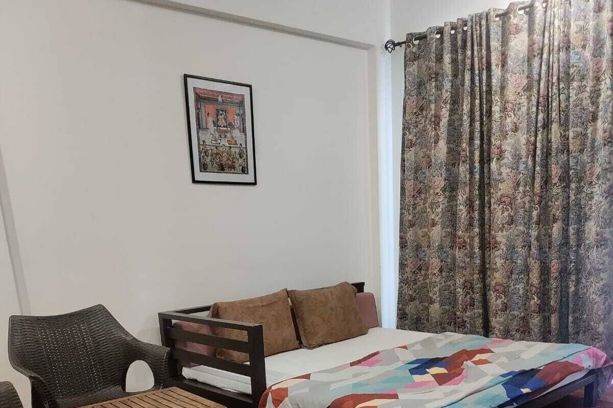 Um quarto pequeno e acolhedor, com uma cama, uma poltrona, uma mesa pequena e um quadro na parede.