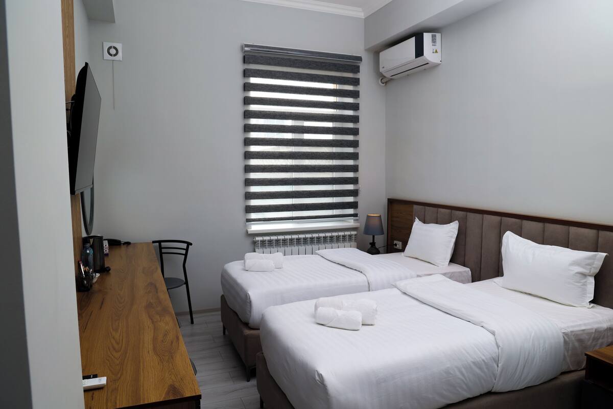 A hotel room with two beds, a wooden desk, a chair, a window with blinds, an air conditioning unit, and a lamp.